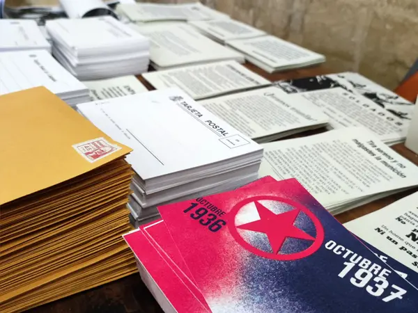 Mesa de trabajo con documentación y material gráfico para asesoramiento a escritores, incluyendo réplicas de tarjetas postales, correspondencia y documentos de la época de la Guerra Civil Española.