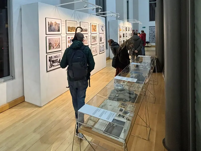 Vista general de la sala de la exposición «50 años de desmemoria», con vitrinas de documentos y un muro de fotografías mientras varios visitantes recorren la muestra.