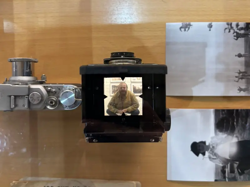 Vitrina con cámaras antiguas y fotografías históricas; en el visor de una cámara de formato medio se refleja Sean Edwards, fotógrafo especializado en cámaras antiguas e históricas.