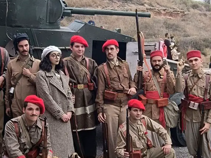 Grupo de recreadores históricos de GRH El Comité con boina roja y uniforme de requetés, junto a una civil con boina blanca, posando frente a un blindado durante la recreación del final de la Batalla del Ebro en Flix (2025).