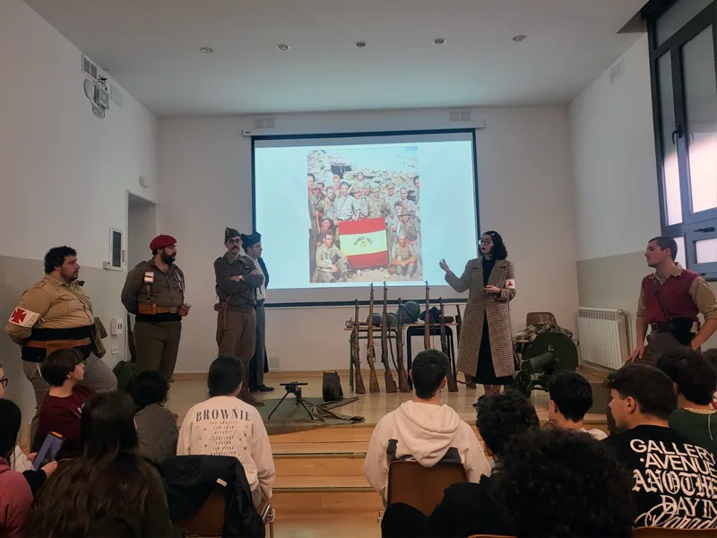 Recreadores de El Comité presentando sus personajes de la Guerra Civil Española ante alumnos de 4.º de la ESO en el salón de actos del Colegio Escolapios de Barbastro, con una fotografía histórica proyectada y armamento original expuesto en el escenario